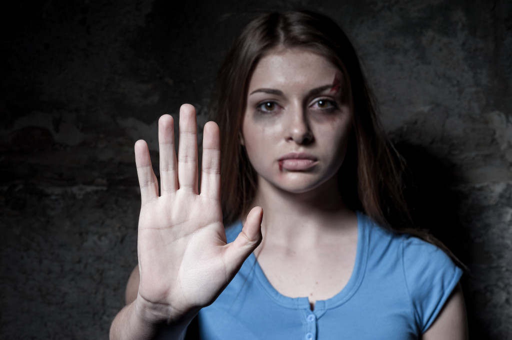 Stop hurting woman! Young beaten up woman looking at camera and stretching out hand while standing against dark wall
