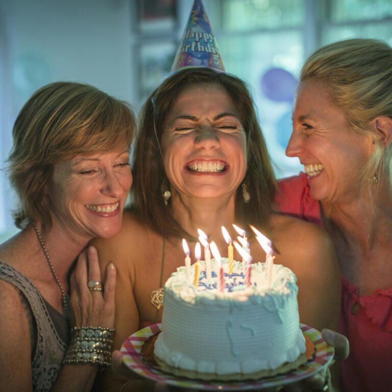 Mature woman holding birthday cake, making wish while two friends look on