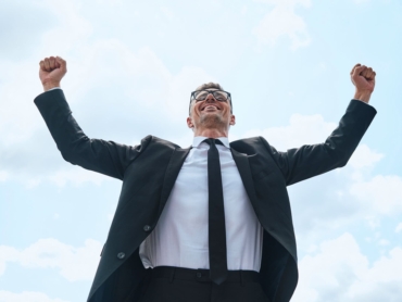 Happy mature man in full suit keeping arms raised while standing in front of blue sky outdoors