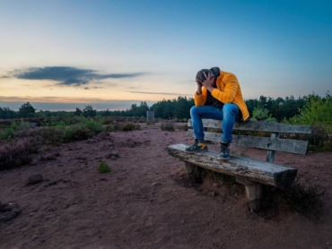 A sad older male clutching his head in his hands on an old wooden bench on a rural field