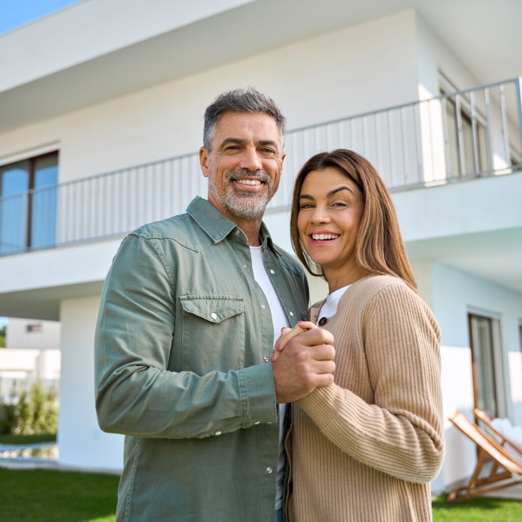 Portrait of happy mature family couple property owners standing outside house.
