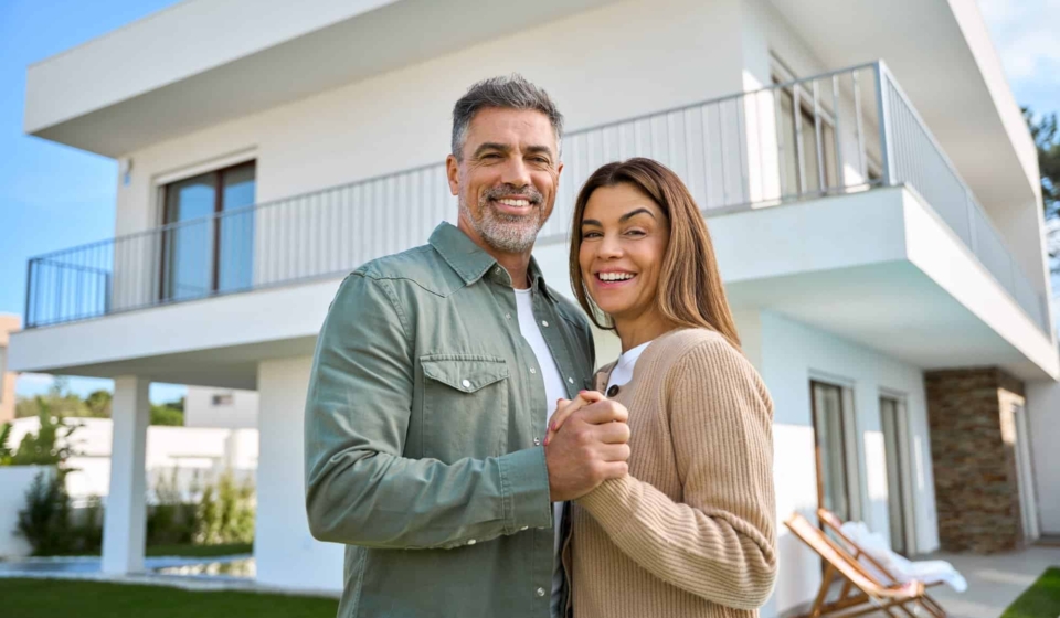 Portrait of happy mature family couple property owners standing outside house.