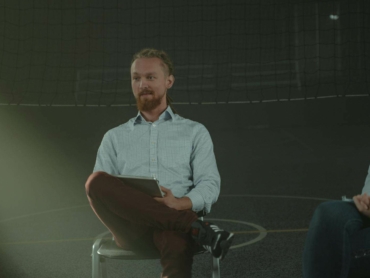 A man holds a meeting in a dimly lit room, engaging with others in a discussion.