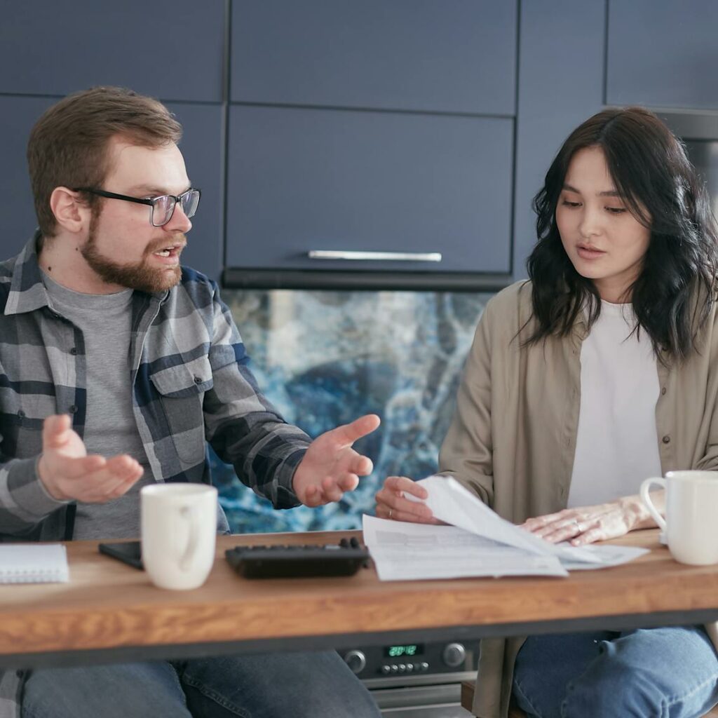 A man and woman discussing financial documents at a kitchen counter, looking concerned.