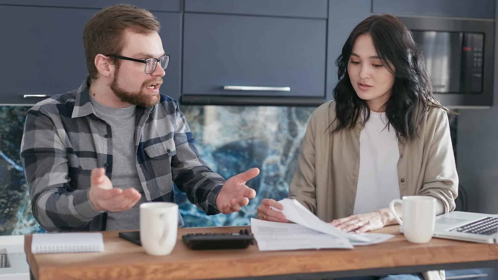 A man and woman discussing financial documents at a kitchen counter, looking concerned.