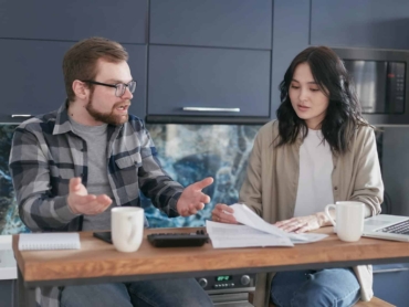A man and woman discussing financial documents at a kitchen counter, looking concerned.