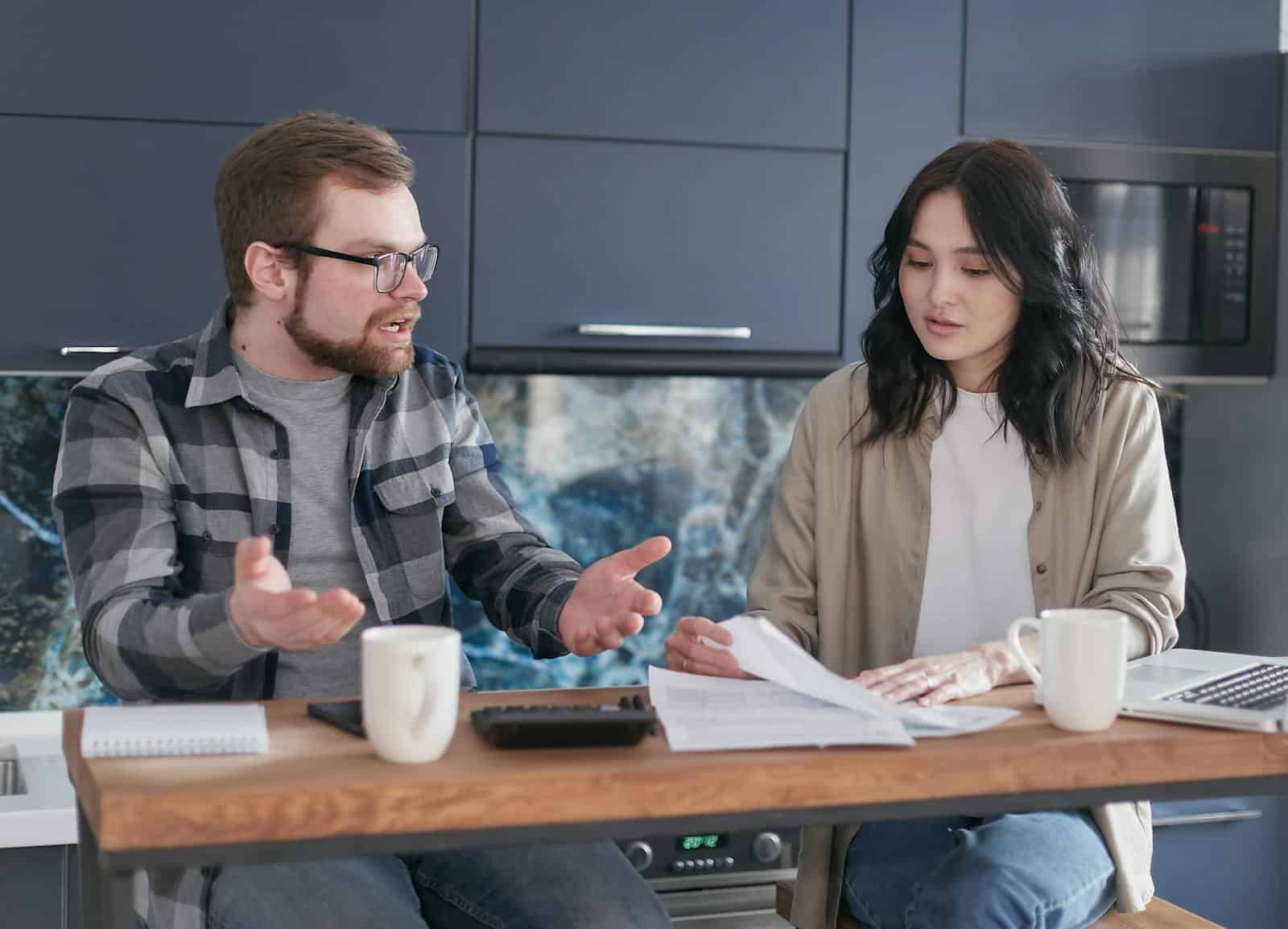 A man and woman discussing financial documents at a kitchen counter, looking concerned.