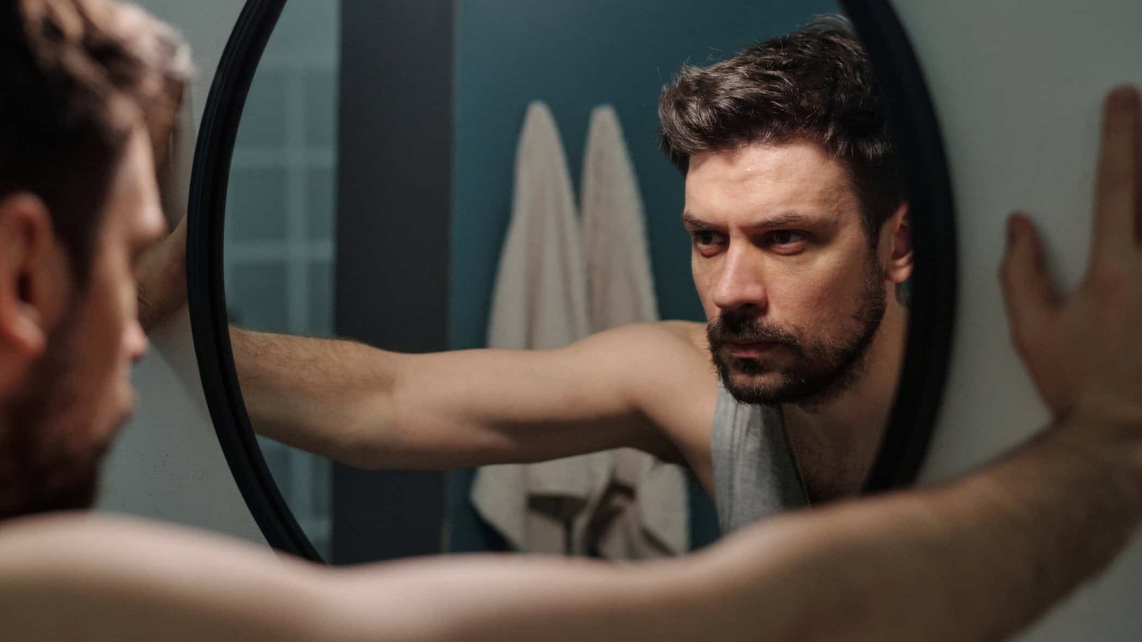 Caucasian young adult man standing in front of mirror gripping frame with both hands reflecting on personal issues showing serious facial expression in bathroom setting