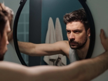 Caucasian young adult man standing in front of mirror gripping frame with both hands reflecting on personal issues showing serious facial expression in bathroom setting