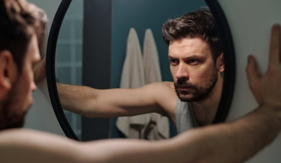 Caucasian young adult man standing in front of mirror gripping frame with both hands reflecting on personal issues showing serious facial expression in bathroom setting