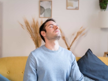 Young bearded male in casual clothes smiling and looking away while resting on couch in cozy living room at home