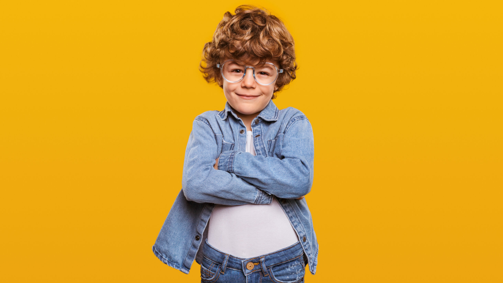 Smart little ginger nerd boy with curly hair wearing denim outfit and round glasses standing with arms crossed against yellow background and looking at camera